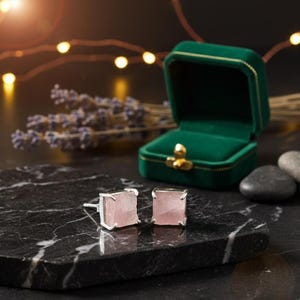 Puede incluir: Pendientes cuadrados de cuarzo rosa con engastes plateados. Los pendientes están sobre una superficie de mármol negro, junto a un joyero de terciopelo verde abierto. Lavanda seca y luces de cadena en el fondo.