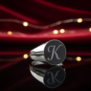 Könnte beinhalten: Ein silberner Siegelring mit einer kreisförmigen schwarzen Einlage auf der Vorderseite. Der Buchstabe K ist in einer kursiven Schriftart in der Mitte der schwarzen Einlage eingraviert. Der Ring hat eine polierte Oberfläche und spiegelt sich.