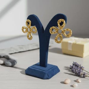 May include: Gold-coloured flower-shaped earrings with a textured finish, displayed on a dark blue velvet earring stand. The earrings have a central white stone. A gift box and lavender sprigs are in the background.