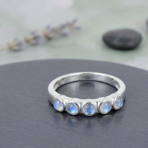 May include: A silver ring featuring five round, cabochon-cut moonstones set in a row along the front of the band. The metal band is stamped with 925, indicating sterling silver composition. The stones exhibit a soft, translucent blue sheen.