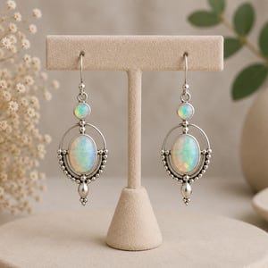 May include: A pair of silver drop earrings featuring oval-shaped opals and smaller round opals. The metalwork includes a circular frame with beaded detailing and a pointed drop at the bottom. The earrings hang from simple silver ear wires.