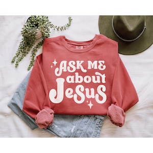 May include: A red long-sleeved sweatshirt with the text "ASK ME about Jesus" in white retro-style lettering. The sweatshirt has a distressed look and features small white stars.