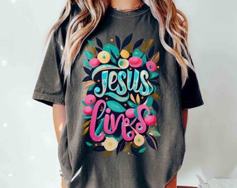 May include: Dark gray t-shirt with the words "Jesus Lives" in a colorful floral design. The text is in a script font, with the word "Jesus" in blue and "Lives" in pink. The floral design includes pink, blue, and yellow flowers.