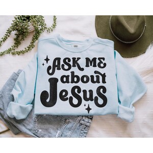 May include: A light blue sweatshirt with the text "Ask me about Jesus" in black retro-style lettering. The text is surrounded by small sparkling stars.