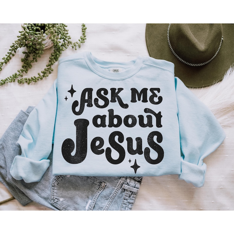 May include: A light blue sweatshirt with the text "Ask me about Jesus" in black retro-style lettering. The text is surrounded by small sparkling stars.
