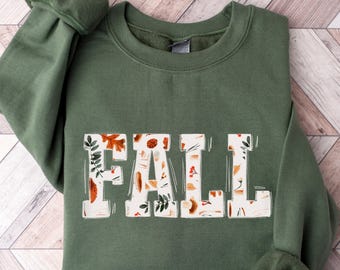 Könnte beinhalten: Ein grüner Pullover mit einem weißen "FALL"-Motiv. Die Buchstaben sind mit einem Muster aus Blättern, Eicheln und Beeren gefüllt.