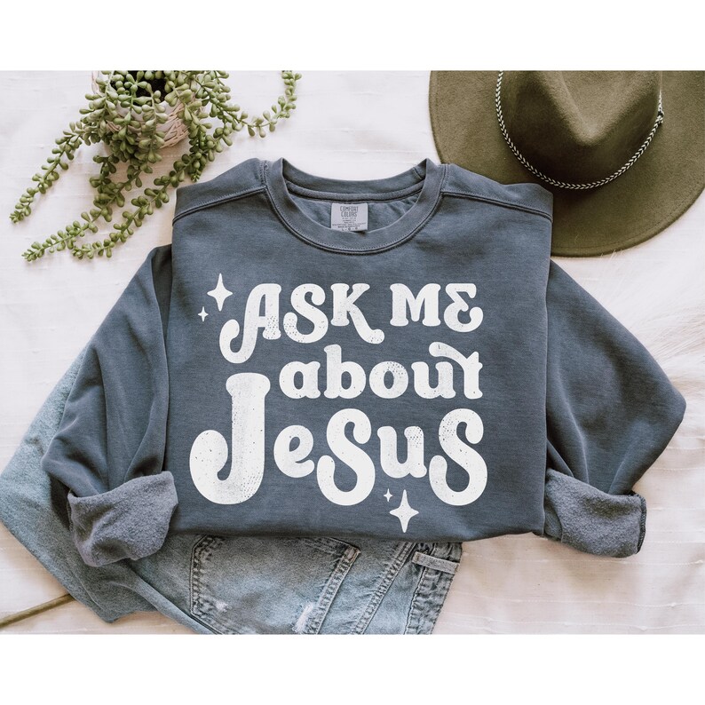 May include: A dark blue sweatshirt with the text "ASK ME about JESUS" in white retro-style lettering. The sweatshirt has a distressed look and features small white stars.