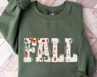 Könnte beinhalten: Ein grüner Pullover mit einer weißen Grafik, auf der in großen Buchstaben "FALL" steht. Die Grafik ist mit einem Muster aus Blättern, Eicheln und Beeren gefüllt.