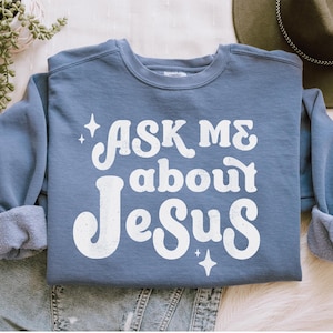 May include: A blue sweatshirt with the text "Ask Me About Jesus" in white retro-style lettering. The text is surrounded by small white stars.