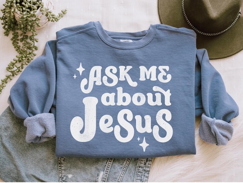 May include: A blue sweatshirt with the text "Ask Me About Jesus" in white retro-style lettering. The text is surrounded by small white stars.