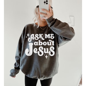 May include: A dark grey sweatshirt with a white graphic that says "Ask Me About Jesus" in a retro font. The graphic also includes two small stars.