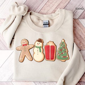 Może przedstawiać: Beżowa bluza z nadrukiem ciasteczek świątecznych: piernikowego ludzika, bałwana, prezentu i choinki. Ciasteczka mają czerwone, zielone i brązowe akcenty. Bluza ma okrągły dekolt i długie rękawy.