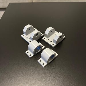 May include: Four light gray metal brackets. Two are rectangular with multiple holes and a raised section with a central hole. The other two are cylindrical with a flat base and two holes for mounting. All have a matte finish.