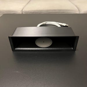 May include: A black rectangular device with a recessed oval-shaped component and a white cable with a USB connector. The device is on a dark surface.