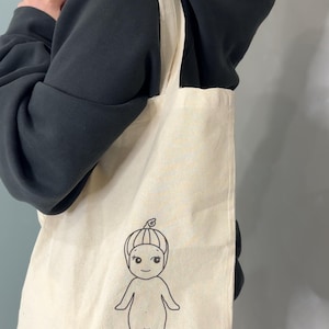 Peut inclure: Une personne portant un sweat &agrave; capuche noir porte un sac fourre-tout blanc avec un dessin au trait noir d'un personnage de dessin anim&eacute;. Le personnage est une personne avec une t&ecirc;te ronde, un petit nez et un sourire. Le personnage porte un chapeau avec une petite feuille sur le dessus.