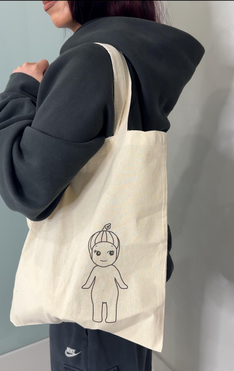 Peut inclure: Une personne portant un sweat &agrave; capuche noir porte un sac fourre-tout blanc avec un dessin au trait noir d'un personnage de dessin anim&eacute;. Le personnage est une personne avec une t&ecirc;te ronde, un petit nez et un sourire. Le personnage porte un chapeau avec une petite feuille sur le dessus.