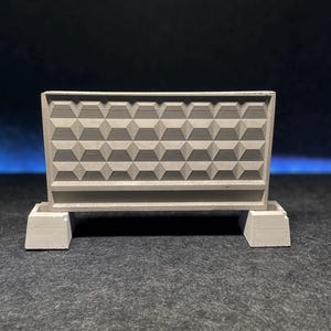 May include: A small, gray concrete barrier with a geometric pattern of raised, square-based pyramids. The barrier is supported by two rectangular bases. The background is a dark gradient of blue and black.