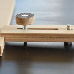 May include: A wooden clamp with a threaded knob and a slot for adjusting the clamp's width. The clamp is made of plywood and has a natural wood finish.
