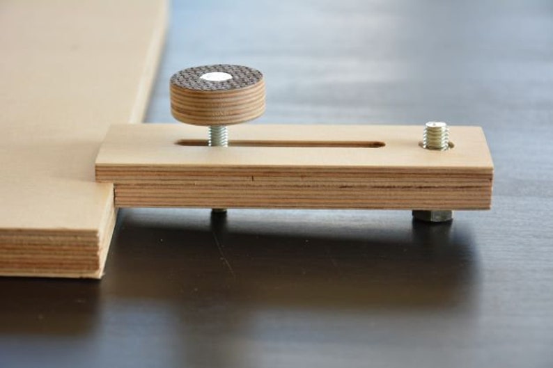 May include: A wooden clamp with a threaded knob and a slot for adjusting the clamp's width. The clamp is made of plywood and has a natural wood finish.