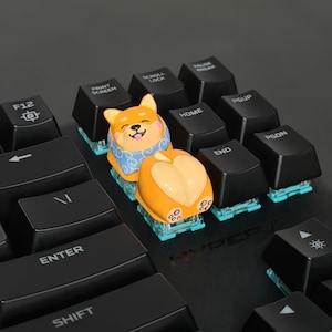 May include: A Shiba Inu dog keycap with a blue and white patterned shirt. The keycap is on a black mechanical keyboard with white lettering. The keys are labeled with functions such as F12, Print Screen, Scroll Lock, Pause Break, Home, PgUp, End, PgDn, Enter, and Shift.