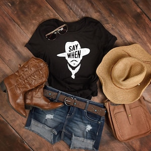 May include: A black t-shirt with the text "SAY WHEN" and a cowboy graphic, paired with denim shorts, a brown belt, cowboy boots, a straw hat, sunglasses, and a brown leather purse. The items are arranged on a wooden surface.
