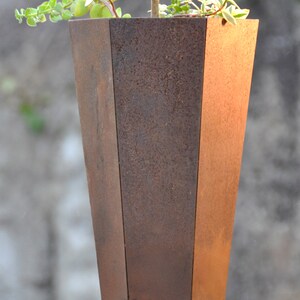 Op de afbeelding: Een roestige metalen plantenbak met een geometrische vorm. De plantenbak is gevuld met groene planten.