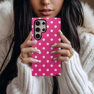 May include: A pink smartphone case with a white polka dot pattern. The case has a camera module with multiple lenses. The phone case is held in front of a person wearing a white sweater.