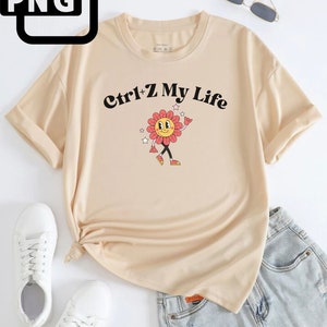 Puede incluir: Una camiseta beige con un gráfico de flor de dibujos animados y el texto "Ctrl+Z My Life" impreso en la parte delantera.