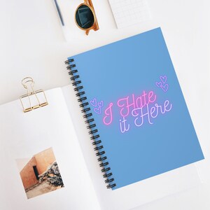 May include: A blue spiral notebook with the text "I Hate it Here" in pink and purple neon lettering. There are small pink hearts around the text.