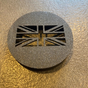 K&ouml;nnte beinhalten: Ein dunkelgraues, kreisf&ouml;rmiges Emblem mit einem ausgeschnittenen Design der Union-Jack-Flagge. Das Emblem hat eine strukturierte Oberfl&auml;che und ist vor einem neutralen Hintergrund platziert. Das Design ist ein Ausdruck des britischen Nationalstolzes.