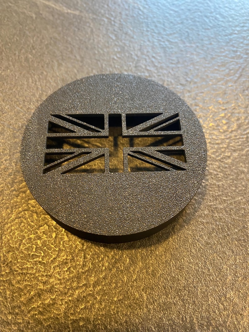 K&ouml;nnte beinhalten: Ein dunkelgraues, kreisf&ouml;rmiges Emblem mit einem ausgeschnittenen Design der Union-Jack-Flagge. Das Emblem hat eine strukturierte Oberfl&auml;che und ist vor einem neutralen Hintergrund platziert. Das Design ist ein Ausdruck des britischen Nationalstolzes.