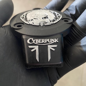 May include: A black circular emblem with a silver-toned metallic inlay featuring the text CYBERPUNK above a stylized graphic symbol. The emblem has a protruding rectangular base and three visible screw holes along its outer edge.