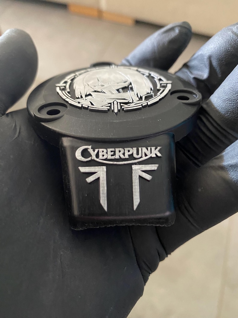 May include: A black circular emblem with a silver-toned metallic inlay featuring the text CYBERPUNK above a stylized graphic symbol. The emblem has a protruding rectangular base and three visible screw holes along its outer edge.