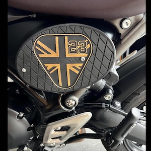 May include: Black motorcycle part with a gold-colored Union Jack flag design and the number 23. The part has a diamond-patterned texture and is mounted on a black motorcycle frame. The seat is brown.
