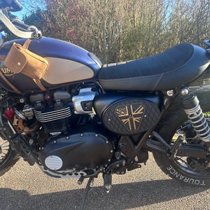 Może przedstawiać: Niebiesko-beżowy motocykl z czarnym siedzeniem i oponami. Mała brązowa sk&oacute;rzana torba jest przymocowana z boku. Motocykl ma czarny panel boczny ze złotym i czarnym wzorem.