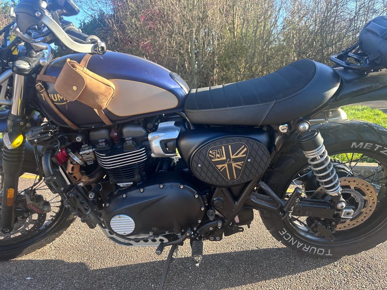 Może przedstawiać: Niebiesko-beżowy motocykl z czarnym siedzeniem i oponami. Mała brązowa sk&oacute;rzana torba jest przymocowana z boku. Motocykl ma czarny panel boczny ze złotym i czarnym wzorem.