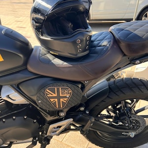 May include: A black motorcycle helmet with a clear visor rests on a brown and black motorcycle seat. The motorcycle has a black frame, a Union Jack emblem, and a black tire. The seat has a diamond pattern.