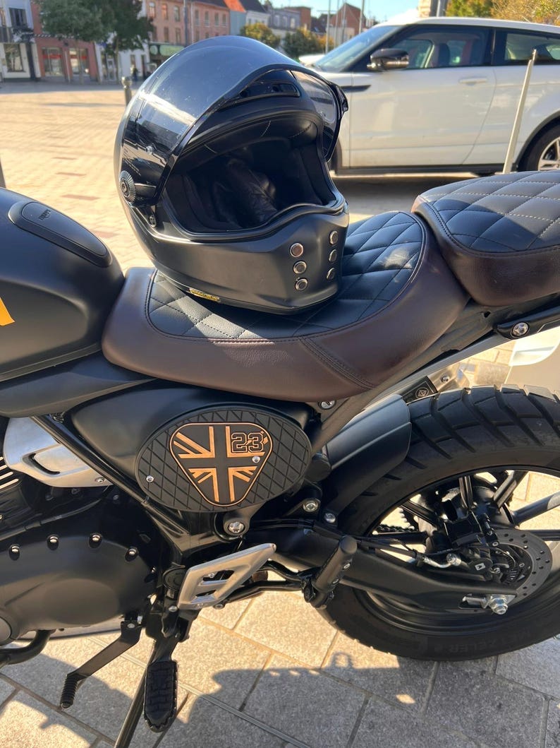 May include: A black motorcycle helmet with a clear visor rests on a brown and black motorcycle seat. The motorcycle has a black frame, a Union Jack emblem, and a black tire. The seat has a diamond pattern.