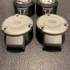 May include: Two pairs of circular motorcycle reservoir covers. The front pair is silver with the text TRIUMPH SINCE 1902 MOTORCYCLES. The rear pair is black with a circular graphic featuring a character and the text CYBERPUNK.