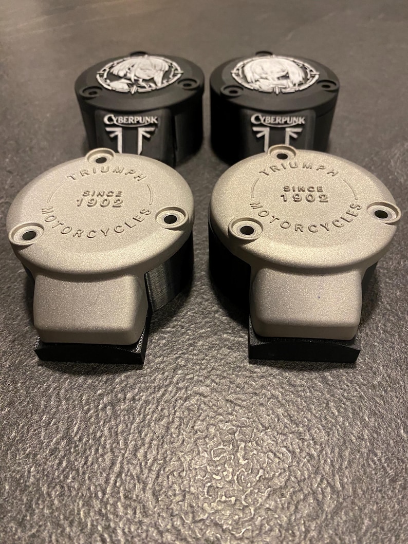 May include: Two pairs of circular motorcycle reservoir covers. The front pair is silver with the text TRIUMPH SINCE 1902 MOTORCYCLES. The rear pair is black with a circular graphic featuring a character and the text CYBERPUNK.