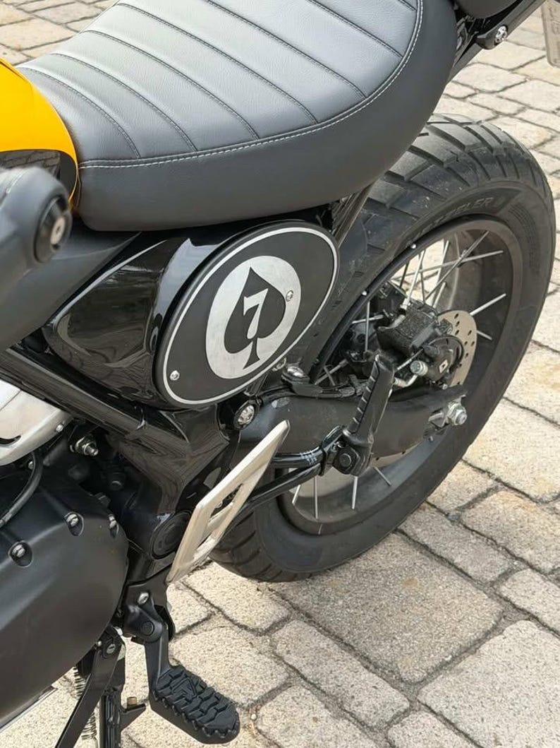 May include: Close-up of a black and yellow motorcycle. The seat is gray with white stitching. A black side panel features a white emblem with a number 7 and a spade symbol. The rear tire is black.