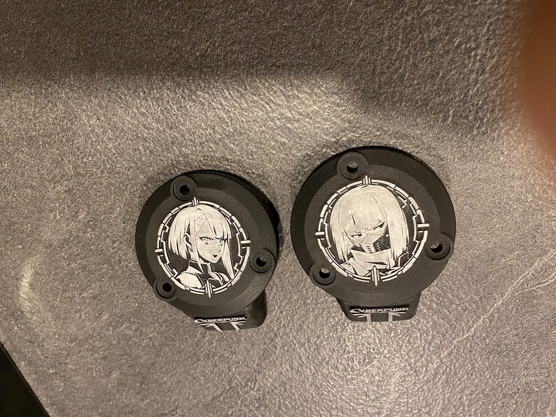 May include: Two circular black metal covers featuring white laser-etched anime-style character portraits within a circular frame. Each cover has two mounting holes and a small rectangular tab at the bottom with the text CYBERPUNK printed in white.
