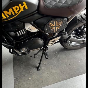 May include: Black motorcycle with yellow "MPH" lettering on the fuel tank. The seat is brown and black diamond-stitched leather. A black side panel features a gold Union Jack and the number 23. The motorcycle is parked on a gray surface.