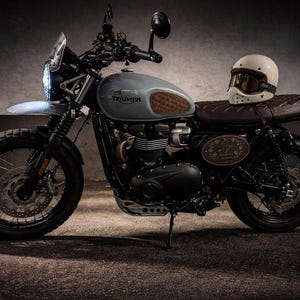 Może przedstawiać: Jasnoniebieski motocykl Triumph z brązowym sk&oacute;rzanym siedzeniem i kremowym kaskiem z goglami. Motocykl ma czarne opony i srebrny przedni błotnik. Słowo "Triumph" jest widoczne na zbiorniku paliwa.