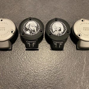 May include: Four circular motorcycle reservoir covers arranged in a row. Two outer covers are silver with the text TRIUMPH SINCE 1902 MOTORCYCLES. Two inner covers are black with white anime-style character graphics and the text CYBERPUNK.
