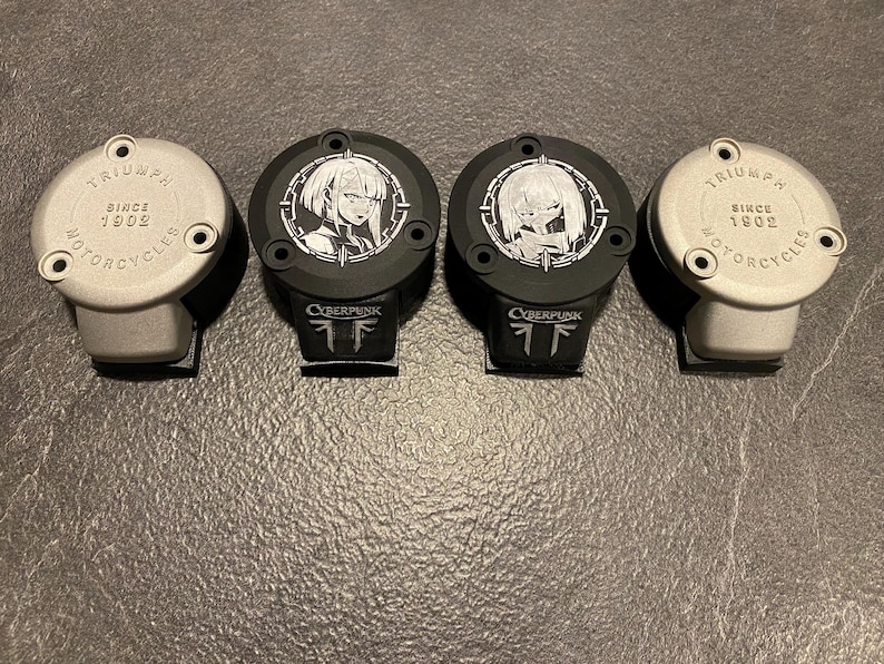 May include: Four circular motorcycle reservoir covers arranged in a row. Two outer covers are silver with the text TRIUMPH SINCE 1902 MOTORCYCLES. Two inner covers are black with white anime-style character graphics and the text CYBERPUNK.