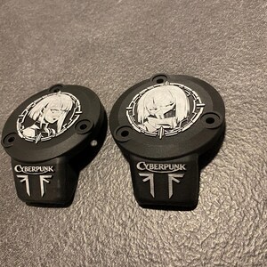 May include: Two matte black circular covers featuring a silver-toned anime-style character portrait within a circular frame. Below the portrait, the word CYBERPUNK is embossed in silver, followed by a stylized logo consisting of two vertical lines.
