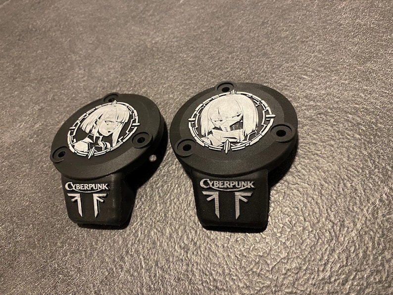 May include: Two matte black circular covers featuring a silver-toned anime-style character portrait within a circular frame. Below the portrait, the word CYBERPUNK is embossed in silver, followed by a stylized logo consisting of two vertical lines.