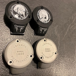 May include: Two black circular motorcycle reservoir covers featuring a white anime character graphic and the text CYBERPUNK, alongside two silver circular reservoir covers embossed with the text TRIUMPH SINCE 1902 MOTORCYCLES, each with three mounting holes.