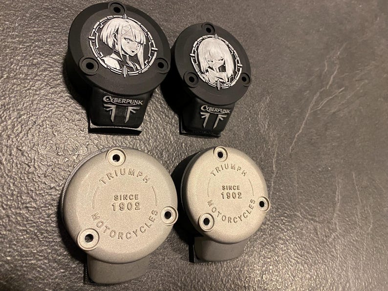 May include: Two black circular motorcycle reservoir covers featuring a white anime character graphic and the text CYBERPUNK, alongside two silver circular reservoir covers embossed with the text TRIUMPH SINCE 1902 MOTORCYCLES, each with three mounting holes.